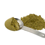 A metal measuring spoon filled with a light olive-green senna leaf powder next to a small pile of the same powder on a white background.