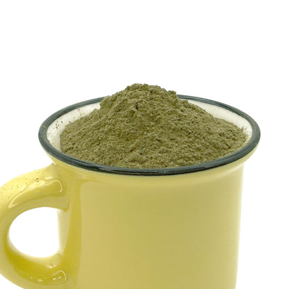 A yellow ceramic mug with a dark rim filled with a mound of green tea leaf powder, shown against a plain white background.