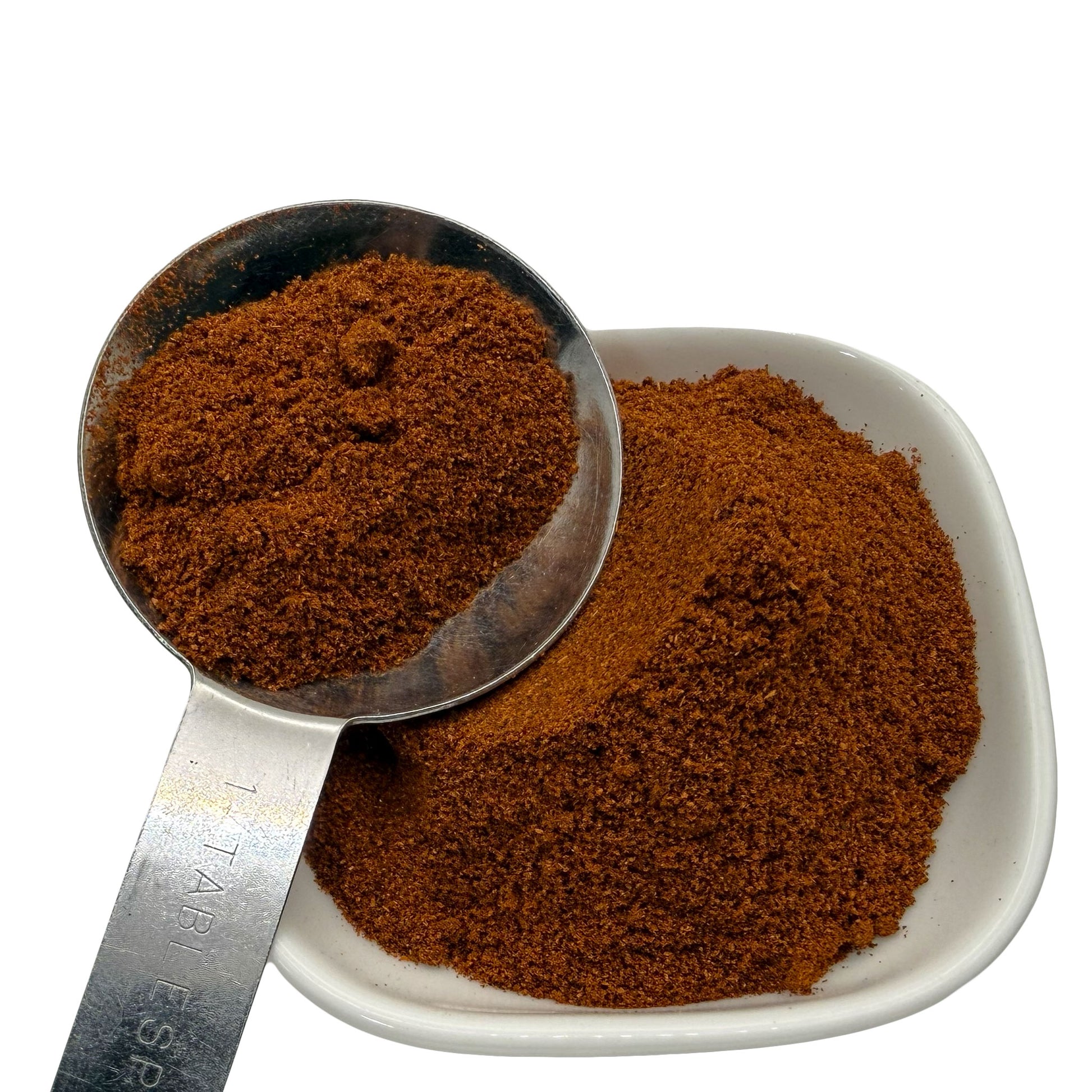 A close-up of a fine reddish brown paprika shown in a metal measuring scoop and a small white square dish against a white background.