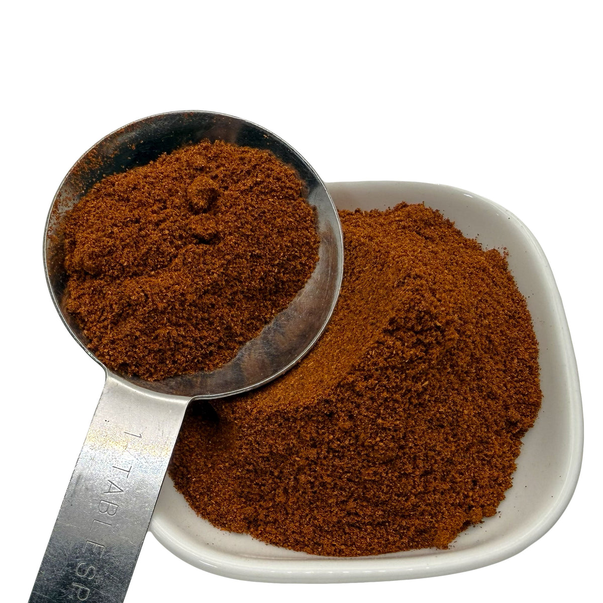 A close-up of a fine reddish brown paprika shown in a metal measuring scoop and a small white square dish against a white background.