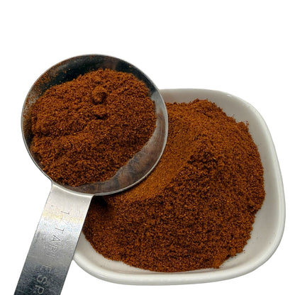 A close-up of a fine reddish brown paprika shown in a metal measuring scoop and a small white square dish against a white background.