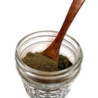 Textured glass jar partially filled with a brownish green witch hazel leaf powder and a wooden spoon
