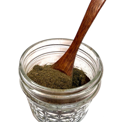 Textured glass jar partially filled with a brownish green witch hazel leaf powder and a wooden spoon